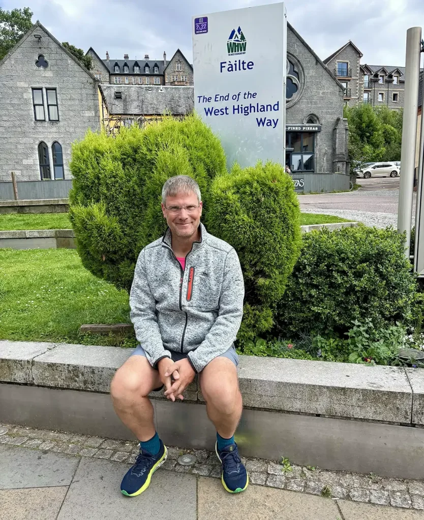 Pater Müller von einer Zypresse auf einer Mauer sitzend, im Hintergrund ein altes Gebäude und ein Schild mit der Aufschrift "The End of the West Highland Way"